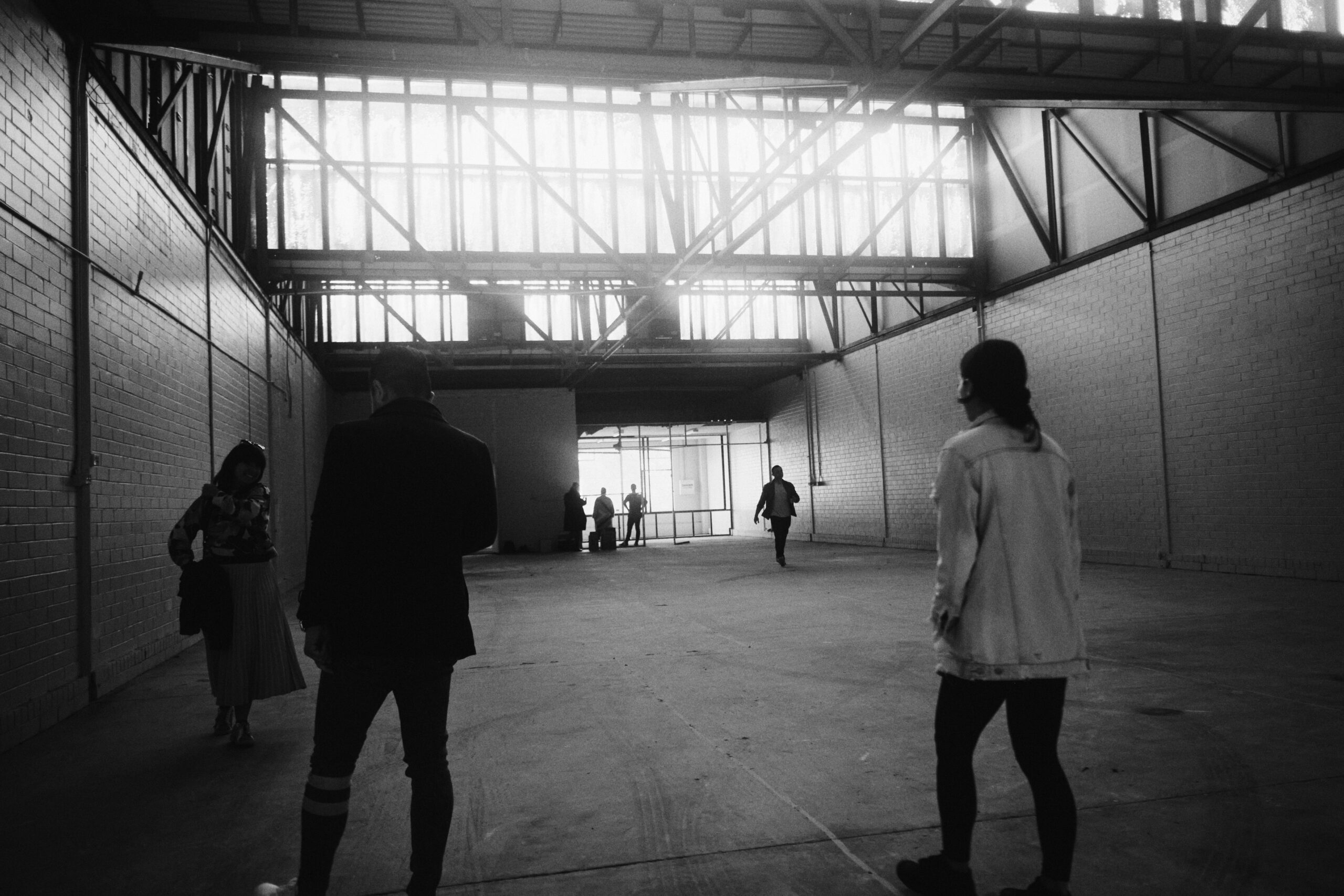 black and white photo of an empty warehouse with saw tooth roof windows and people walking around the space.