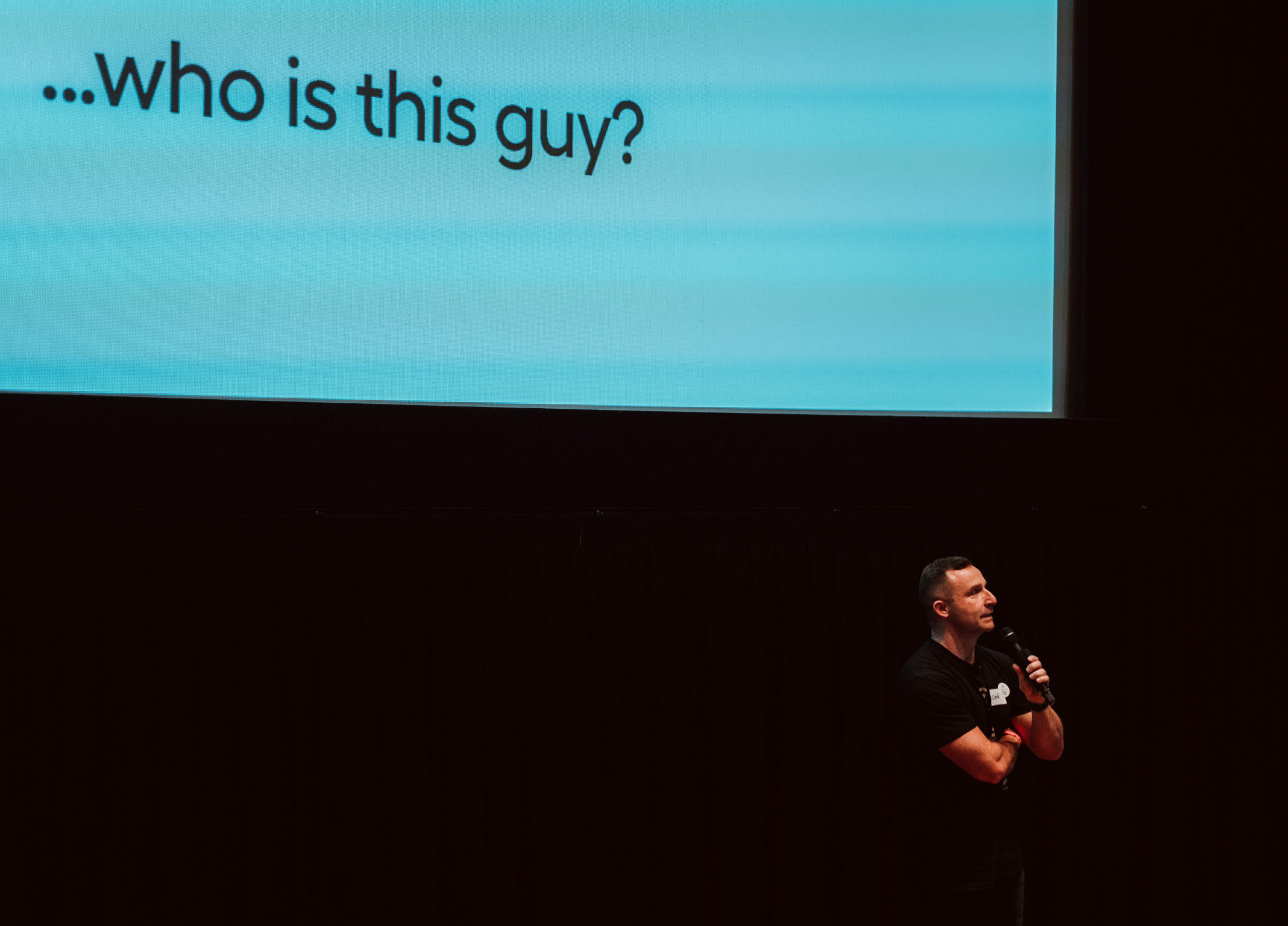 Niaal holding a microphone on stage with screen in the background displaying the text "who is this guy?" on blue.