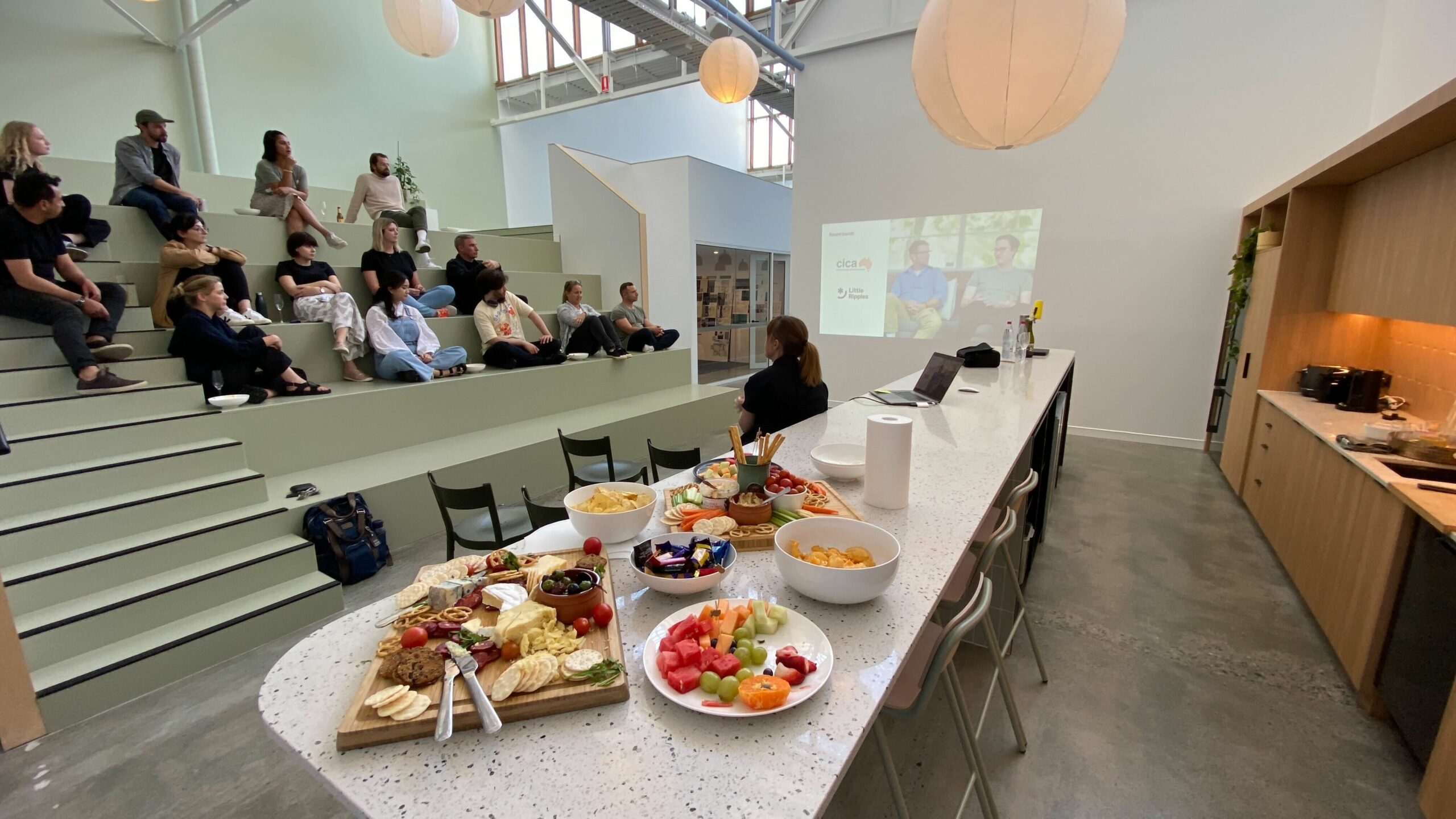 Anthologie office with the team sitting on the green bleachers, a presentation projected on the wall, cheese board in the foreground.